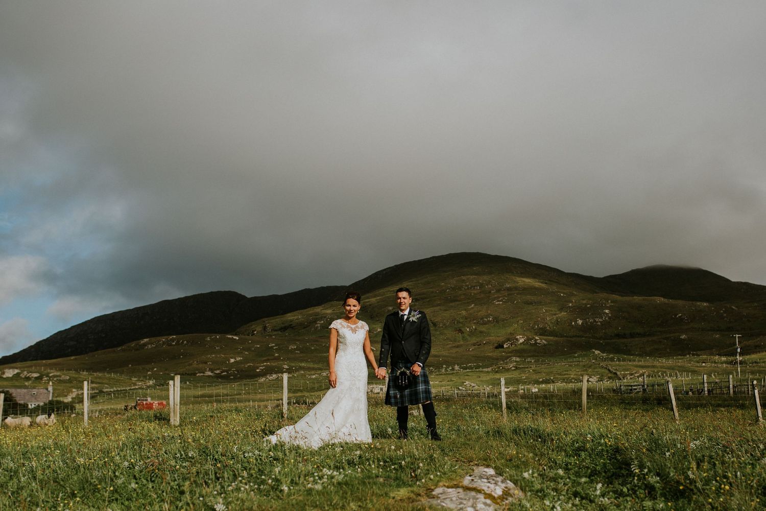 A wedding couple poses in traditional Scottish attire against rolling green hills and rustic fencing.