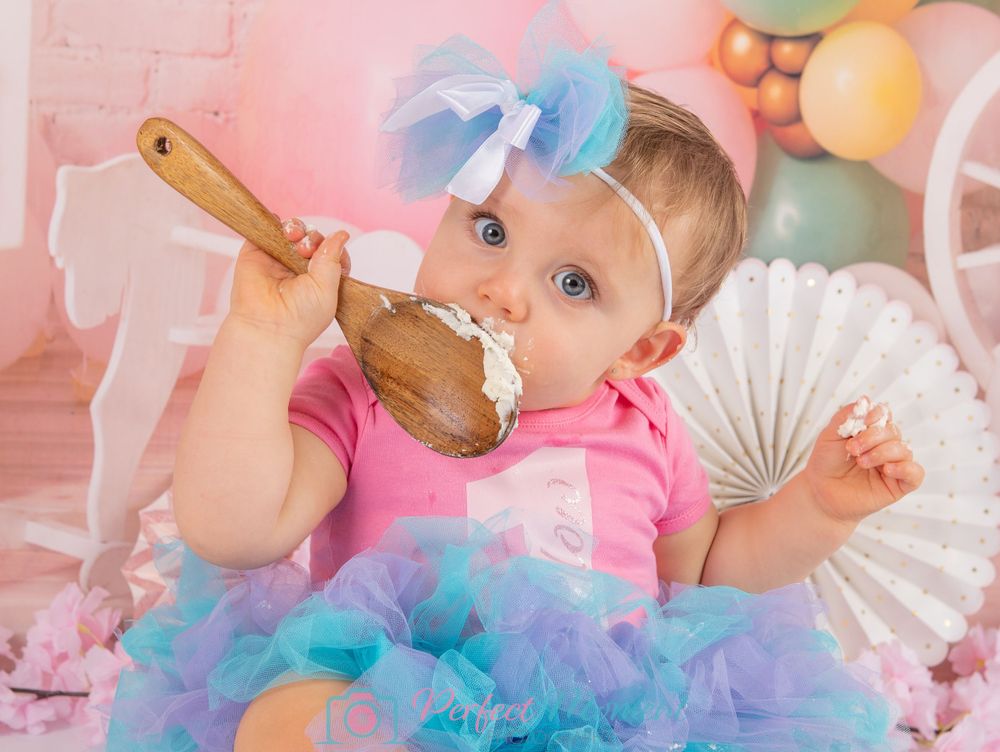 Little girl enjoying her cake smash photo shoot at Solihull studio. She's eating buttercream icing from a wooden spoon, wearing a blue tutu