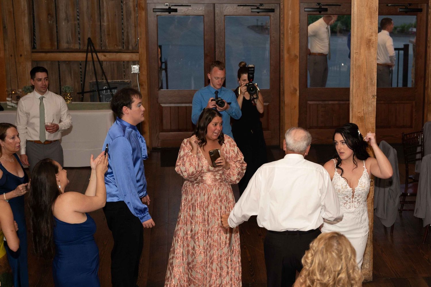 Wedding guests dancing and celebrating at a rustic indoor reception venue with wooden beams and warm lighting.