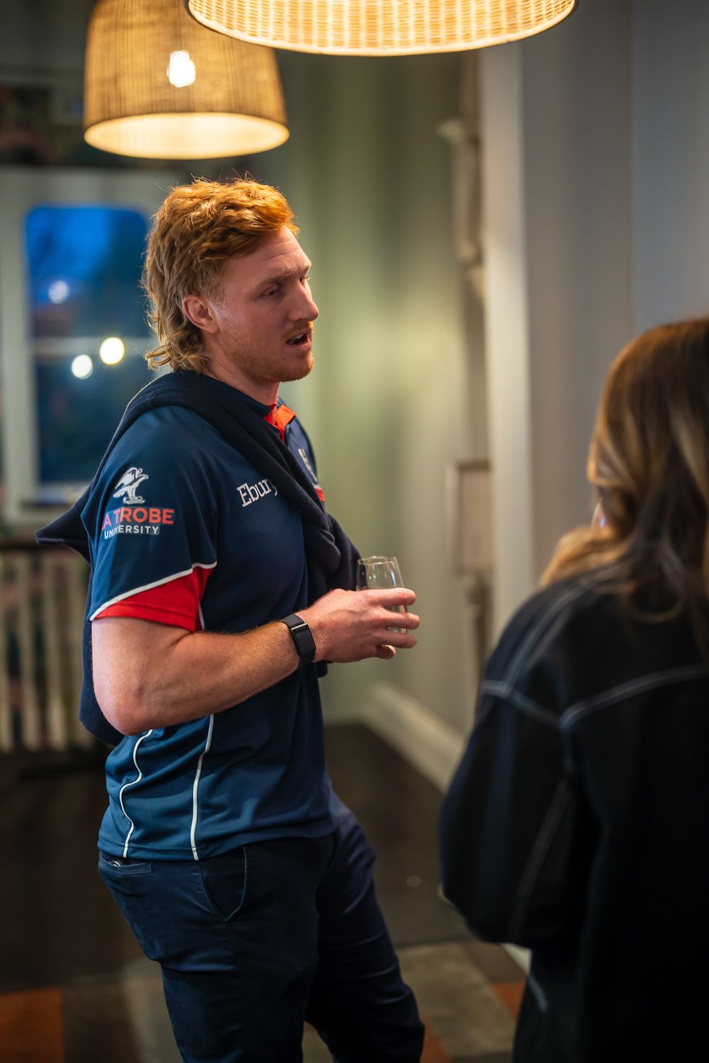 Rugby player in team jersey holding a drink and conversing at an indoor networking event.
