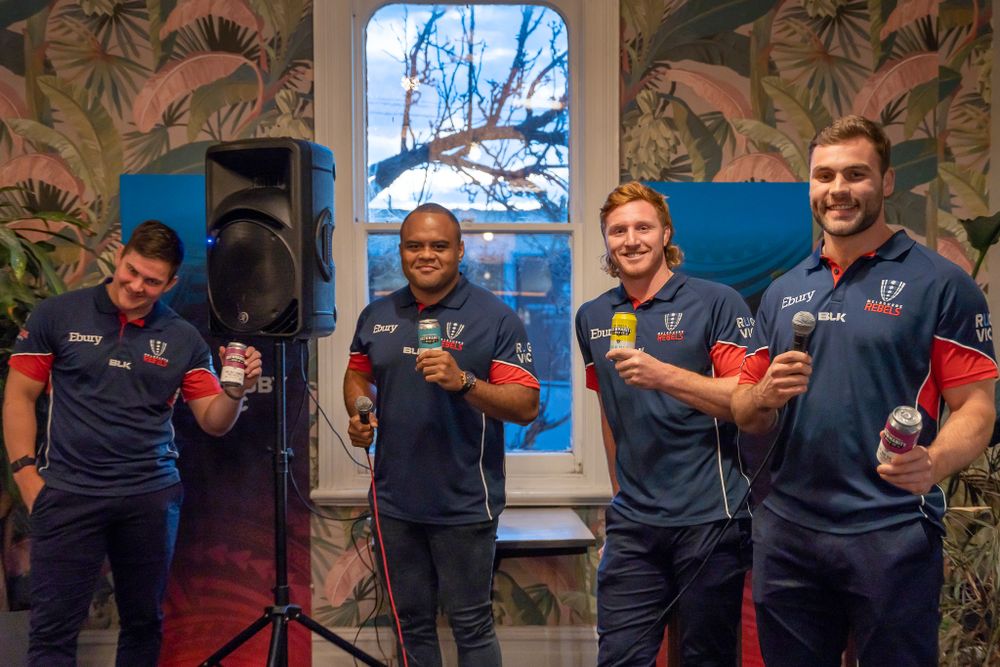 Four men in matching navy sports uniforms smile and pose together at an indoor promotional event.