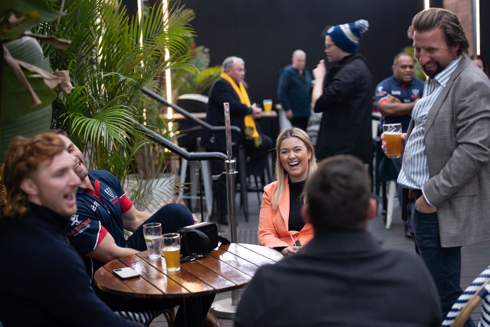 Group of friends laughing and socializing at an outdoor bar patio with drinks.