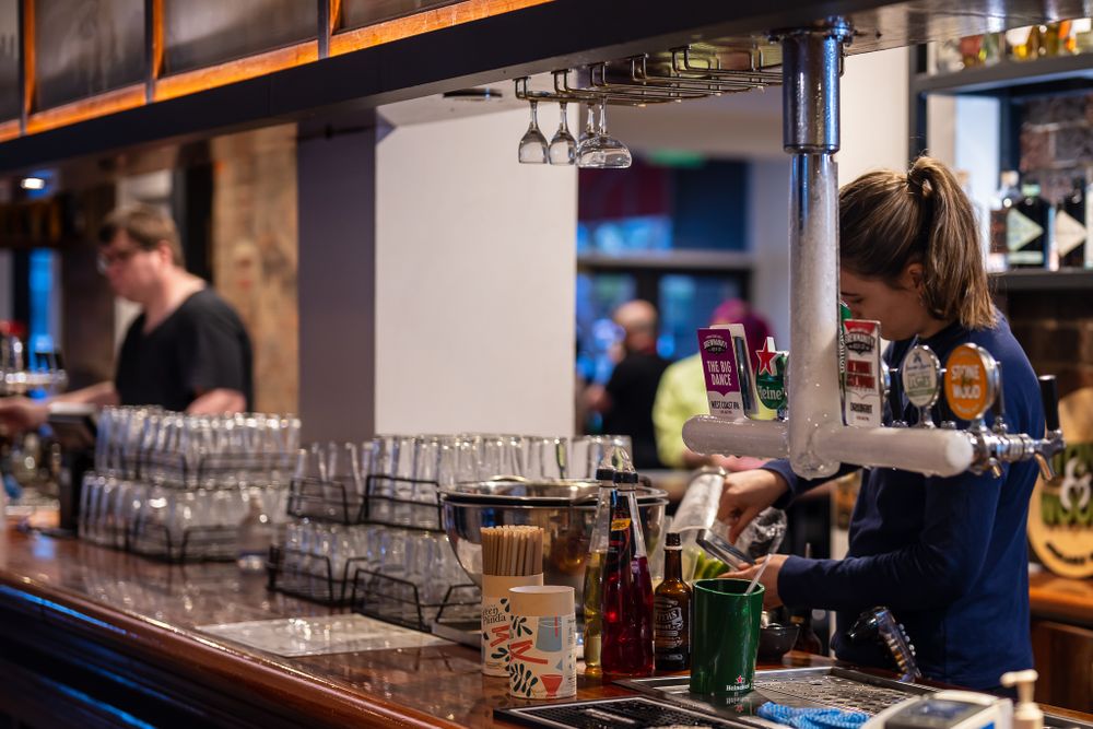 Female bartender pouring drinks at a well-stocked wooden bar counter.
