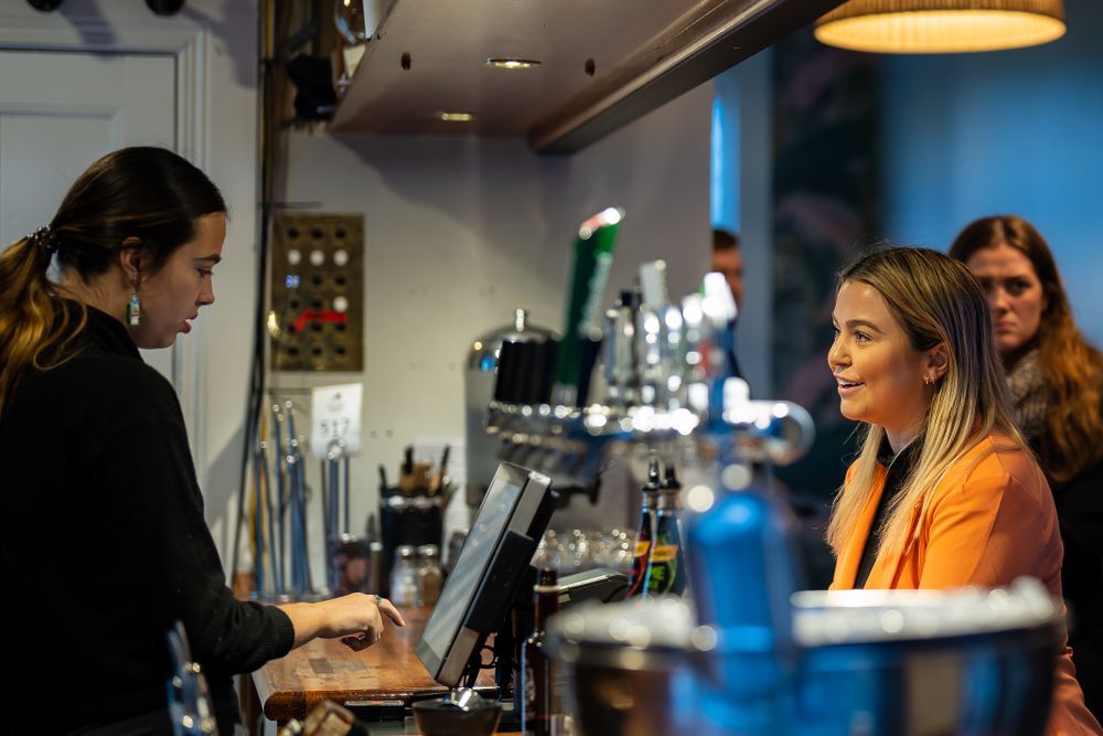 Woman in orange jacket ordering at a modern bar with female bartender.