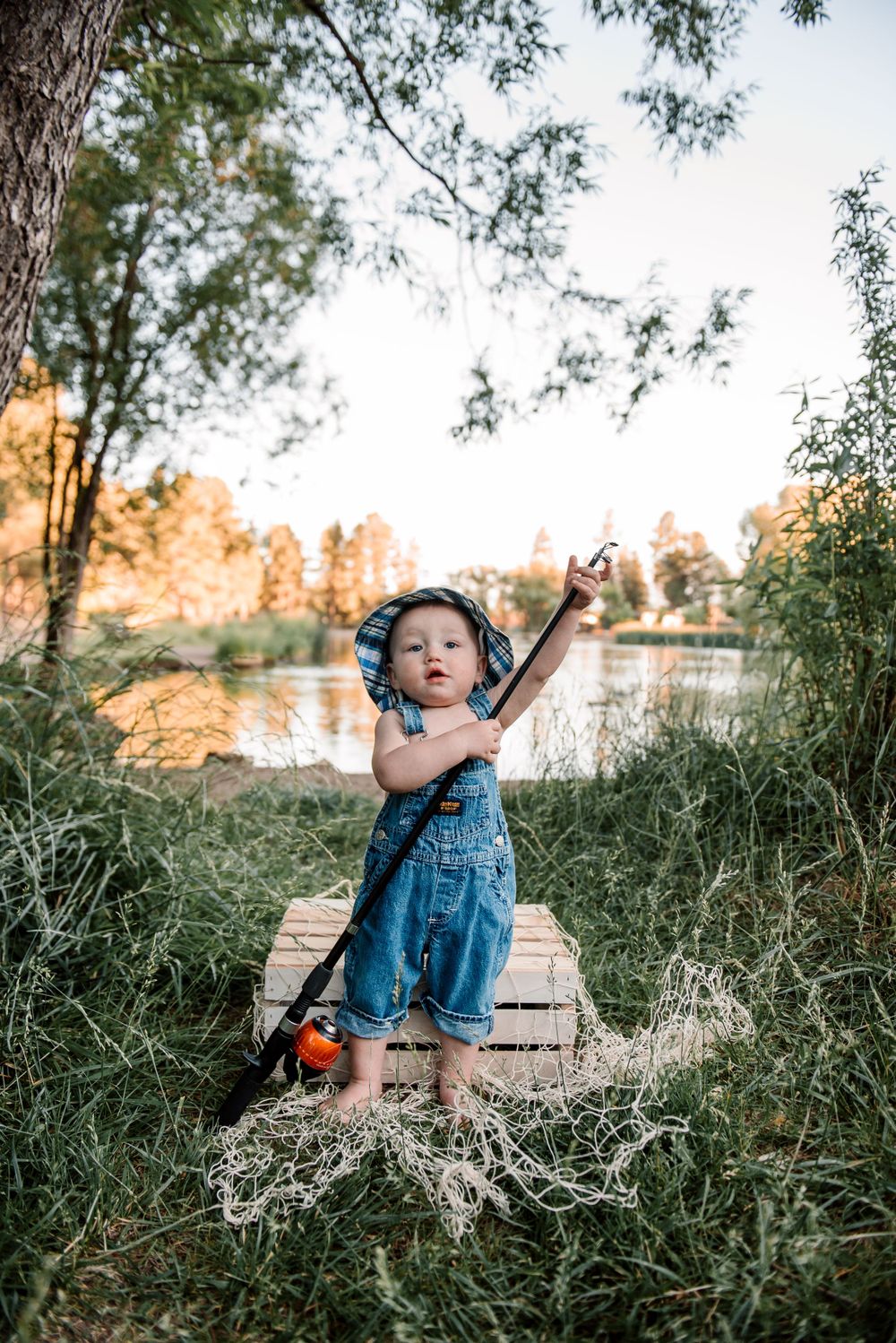 Toddler in denim overalls playing outdoors near lake during golden hour.