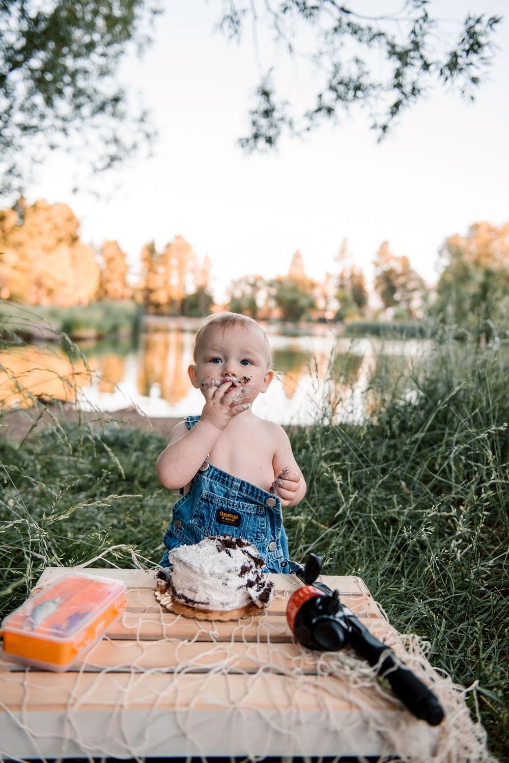 Baby in overalls enjoying messy cake smash photography session outdoors.