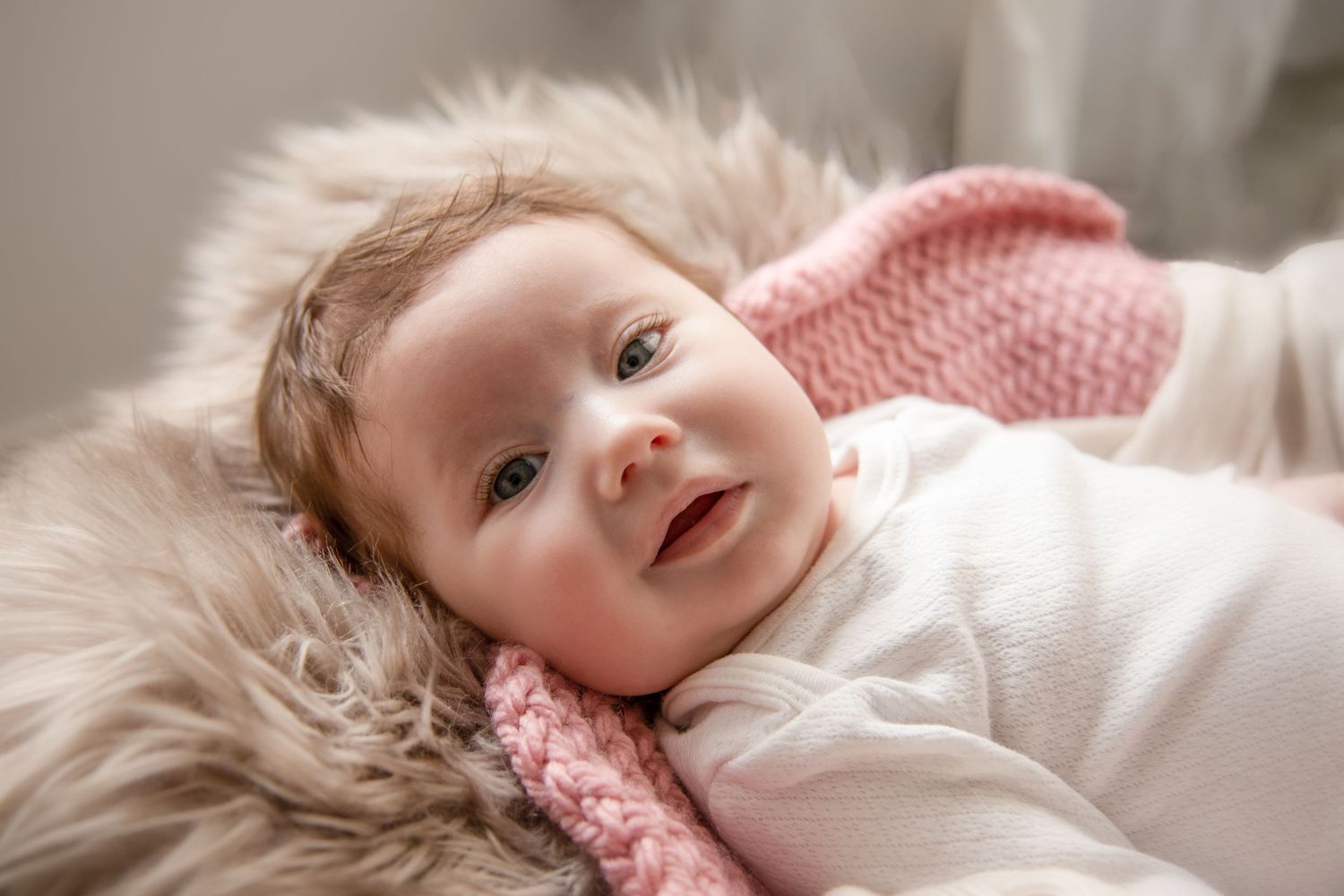 A smiling baby wearing pink lies on soft white fur bedding.