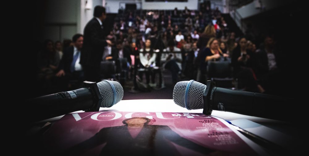 Two microphones on a podium in front of a large audience at a professional conference or speaking event.