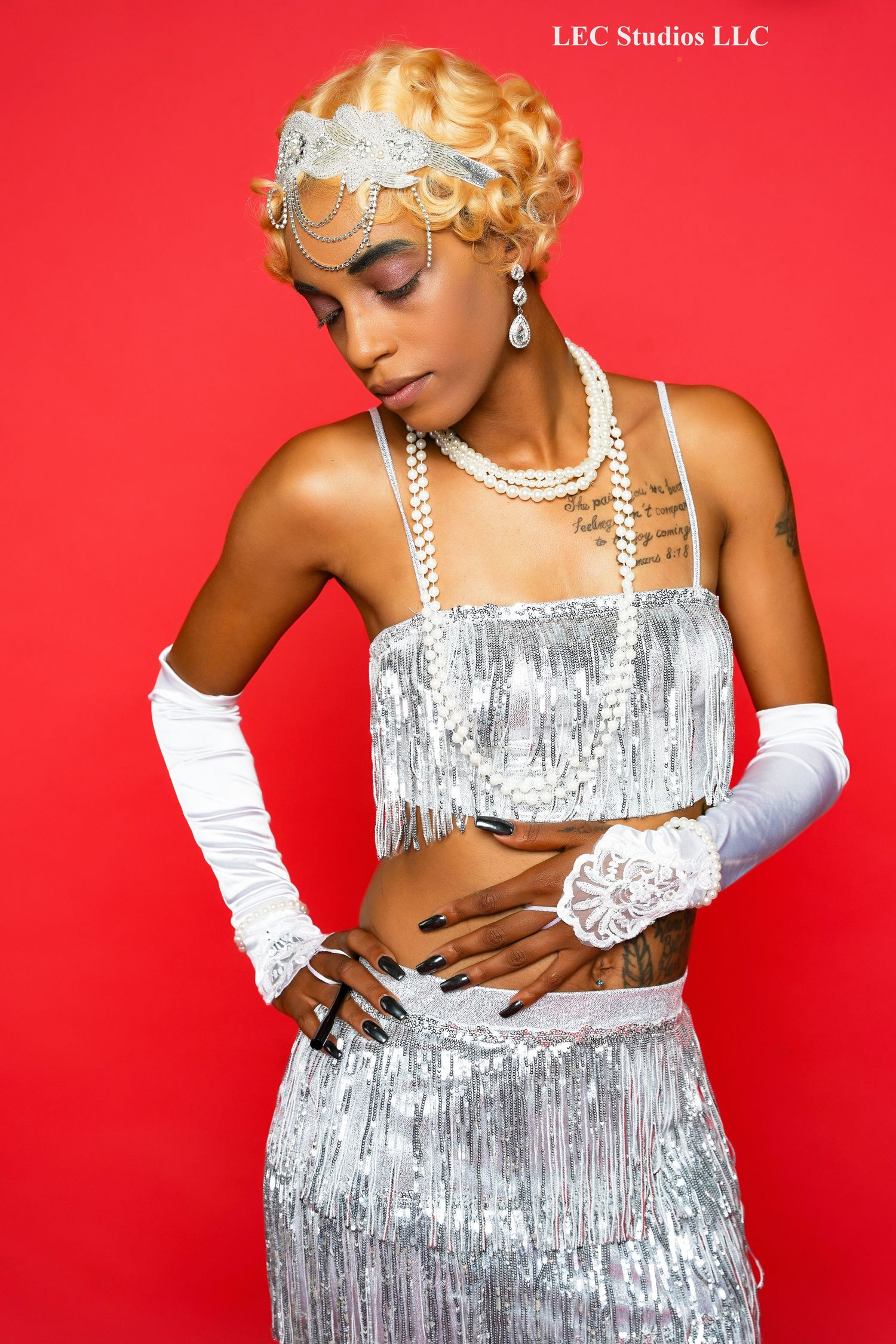 Glamorous 1920s-inspired fashion portrait with silver fringe outfit, pearl necklaces and vintage hair accessories against red backdrop.