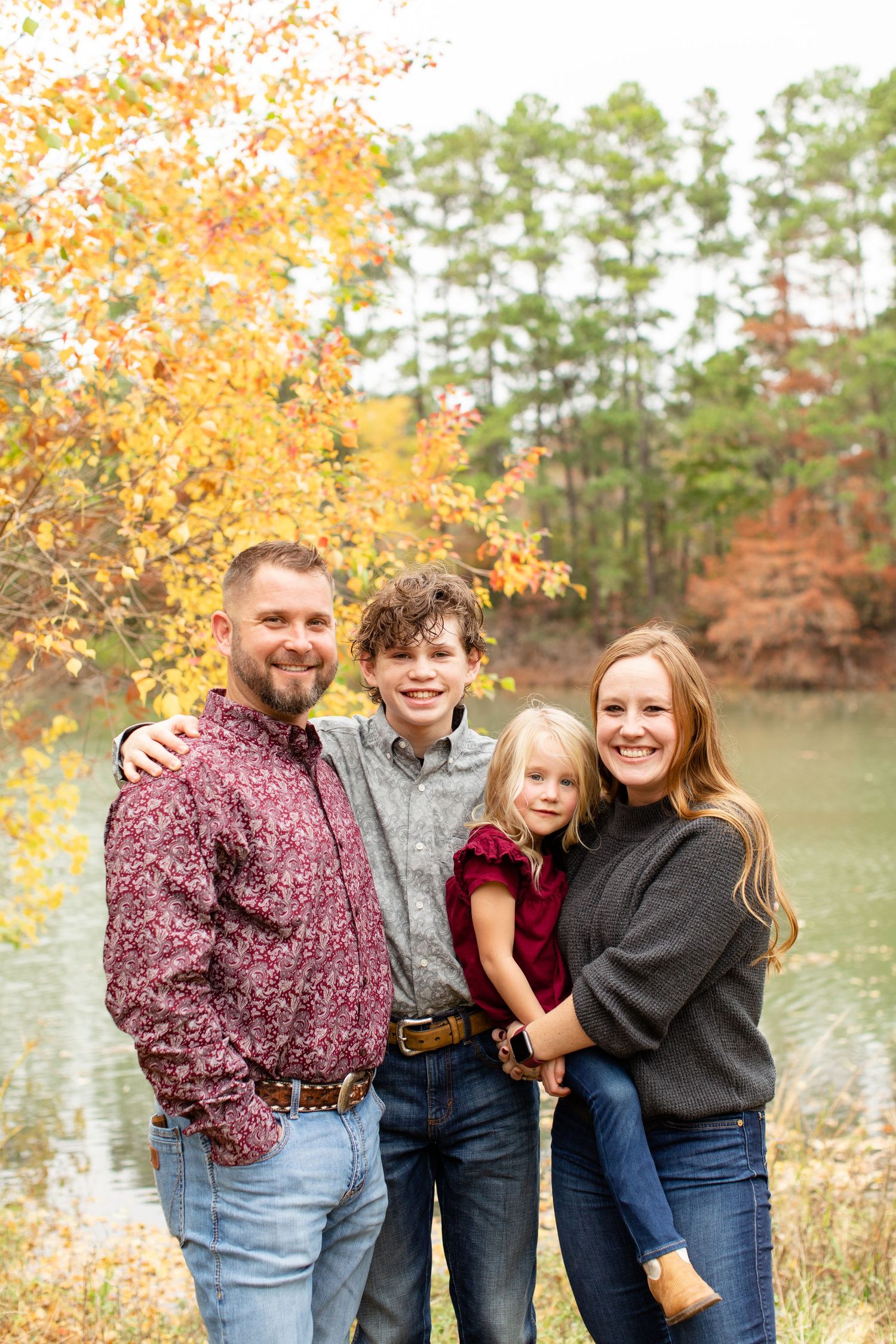 A family portrait by a lake with fall foliage and pine trees in the background.