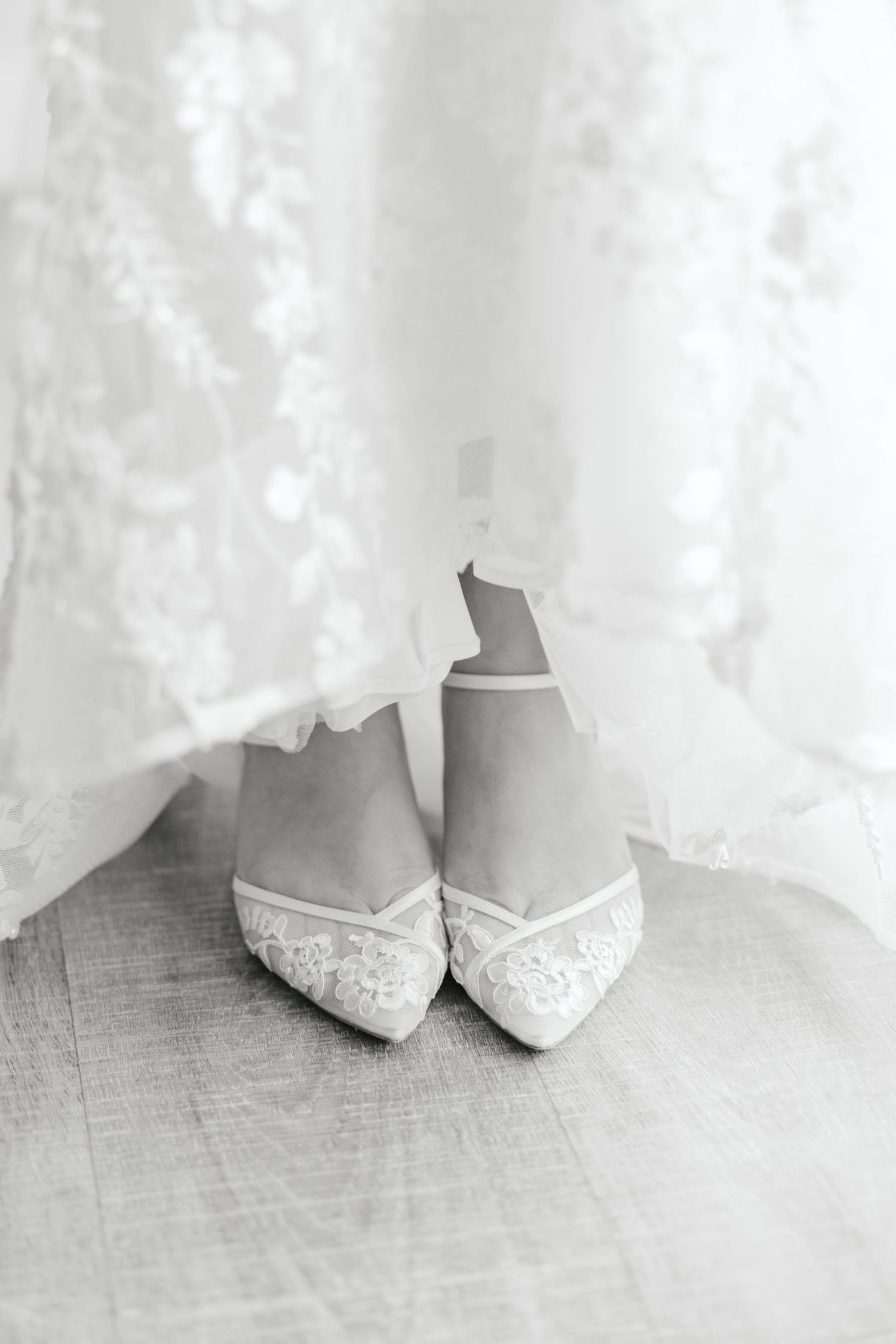 Black and white close-up of elegant lace bridal heels beneath a flowing wedding gown.