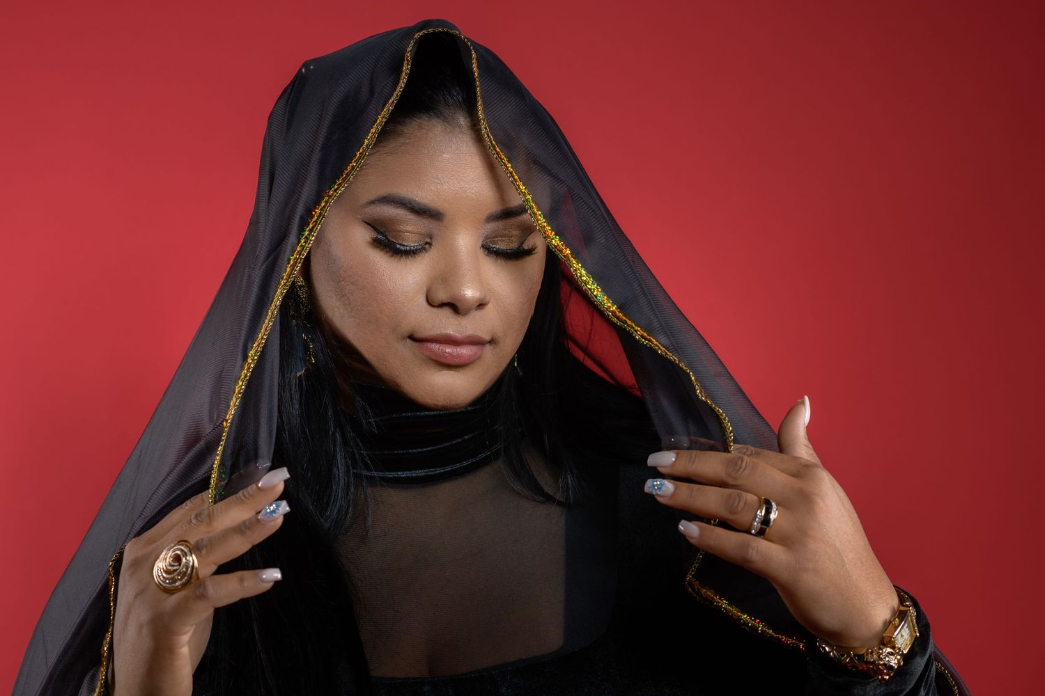 Person wearing black sheer top and headscarf poses elegantly against red background with closed eyes and peaceful expression.