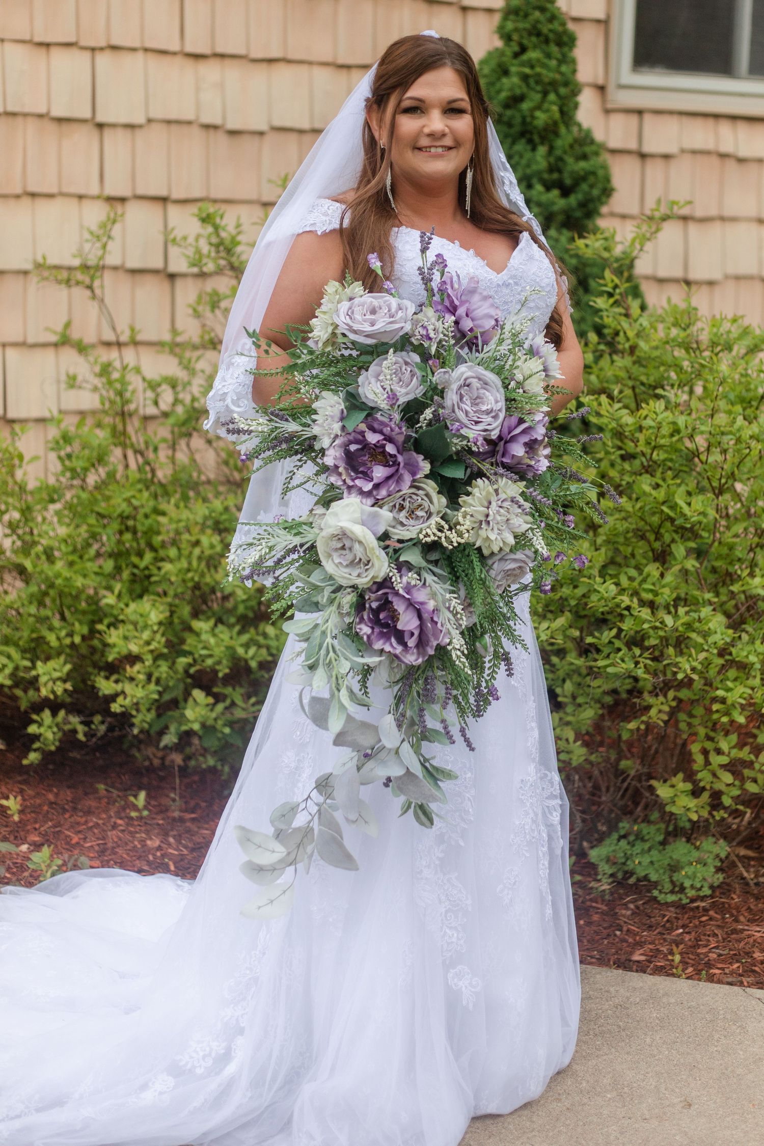 A bride in a white dress holds a stunning purple and white floral bouquet with cascading greenery.