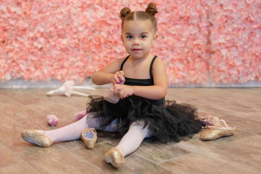 Young ballet dancer in black tutu sitting on wooden floor with pink background.