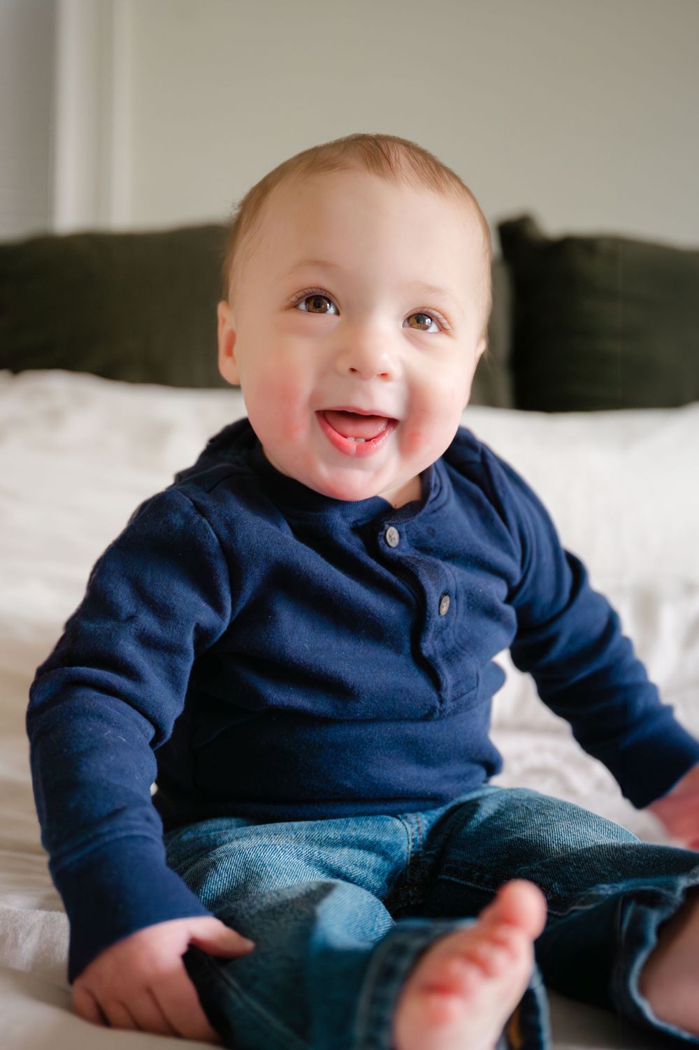 A baby wearing a navy blue sweater sitting on a couch with a bright smile.