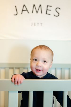 A baby smiling from behind a crib rail with text reading 'JAMES' on the wall.