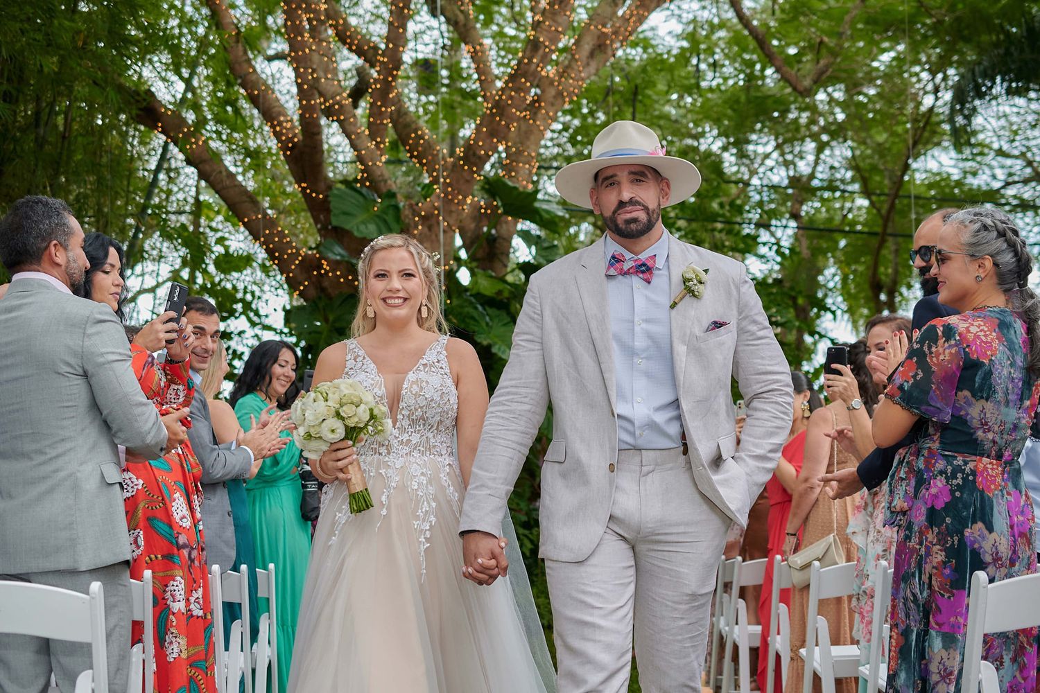 Wedding celebration with guests applauding couple walking down aisle under tree canopy.