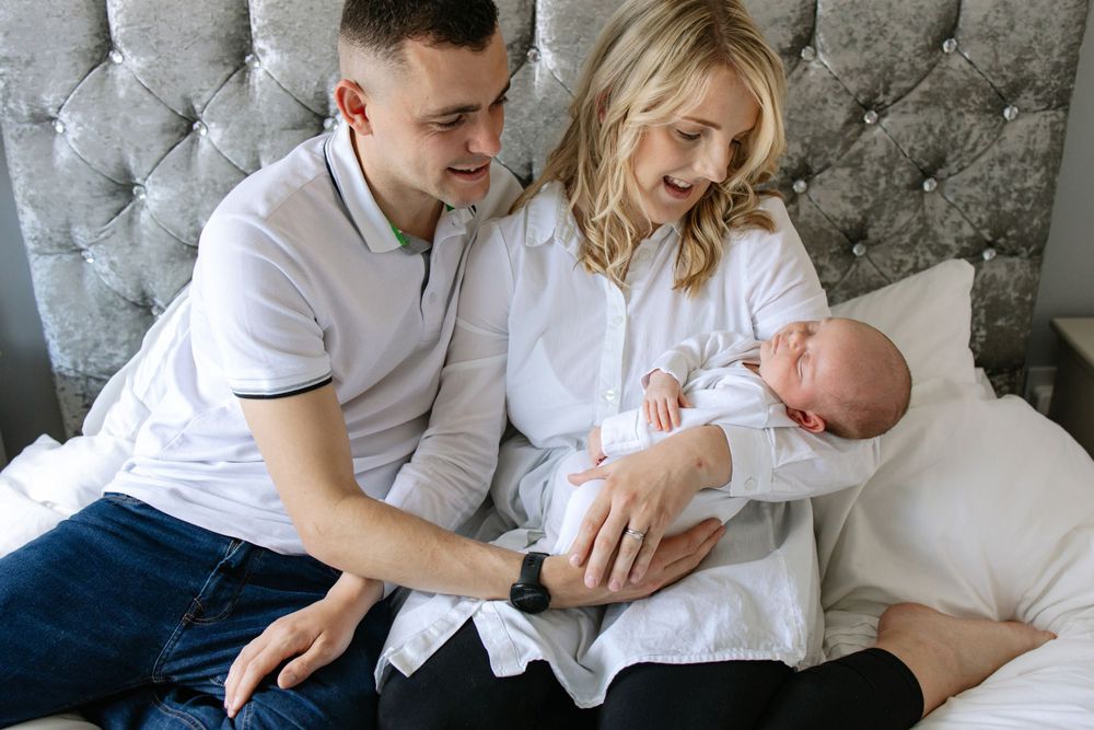 Parents lovingly cradle their newborn baby on a plush gray headboard bed.