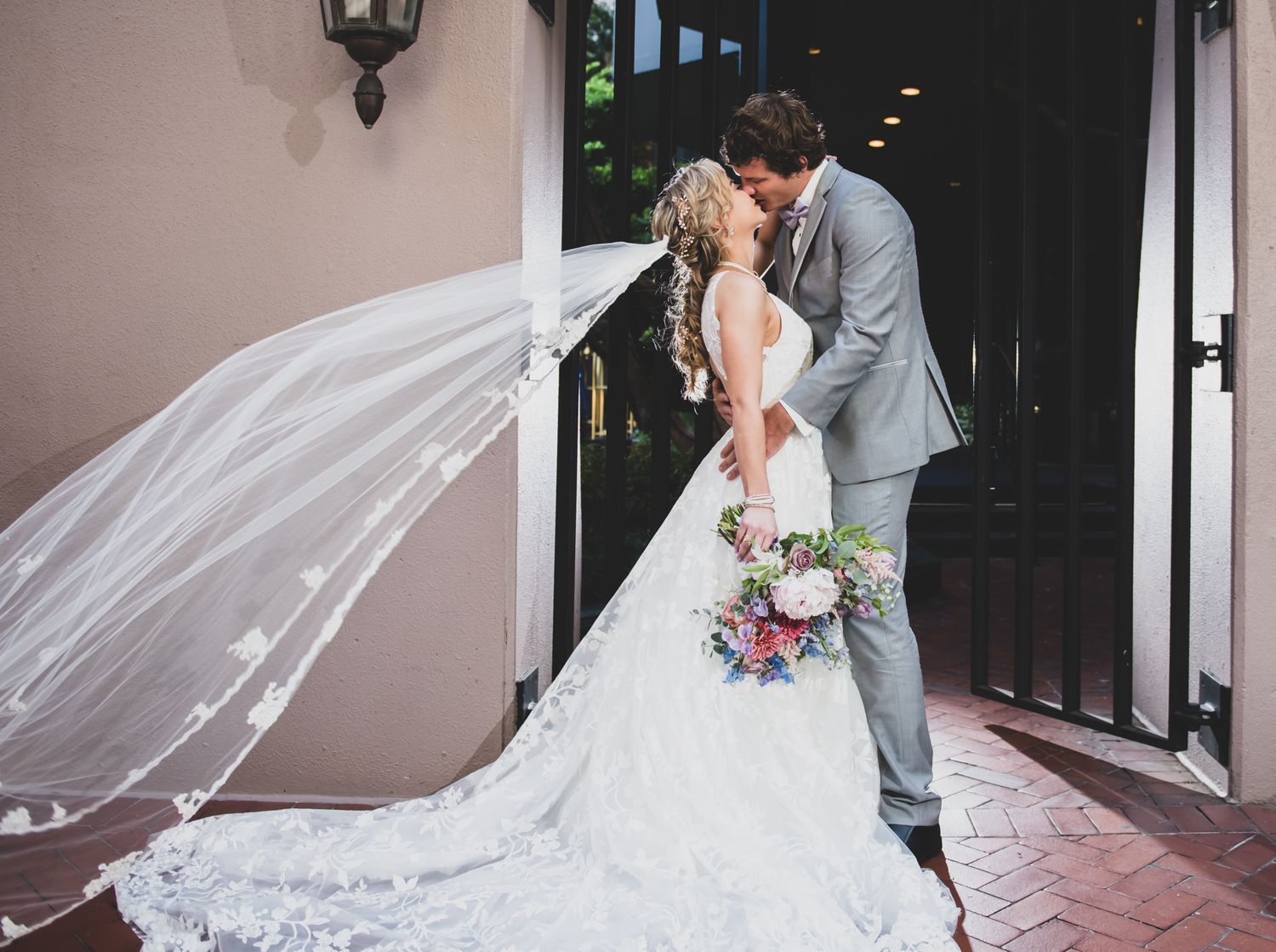 Dramatic wedding photo with flowing veil against pink stucco wall.
