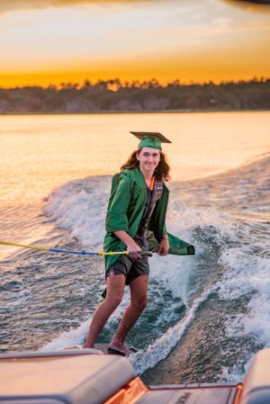 senior wakeboarding on lake with cap and gown during fun candid senior portrait photoshoot Panama City Florida