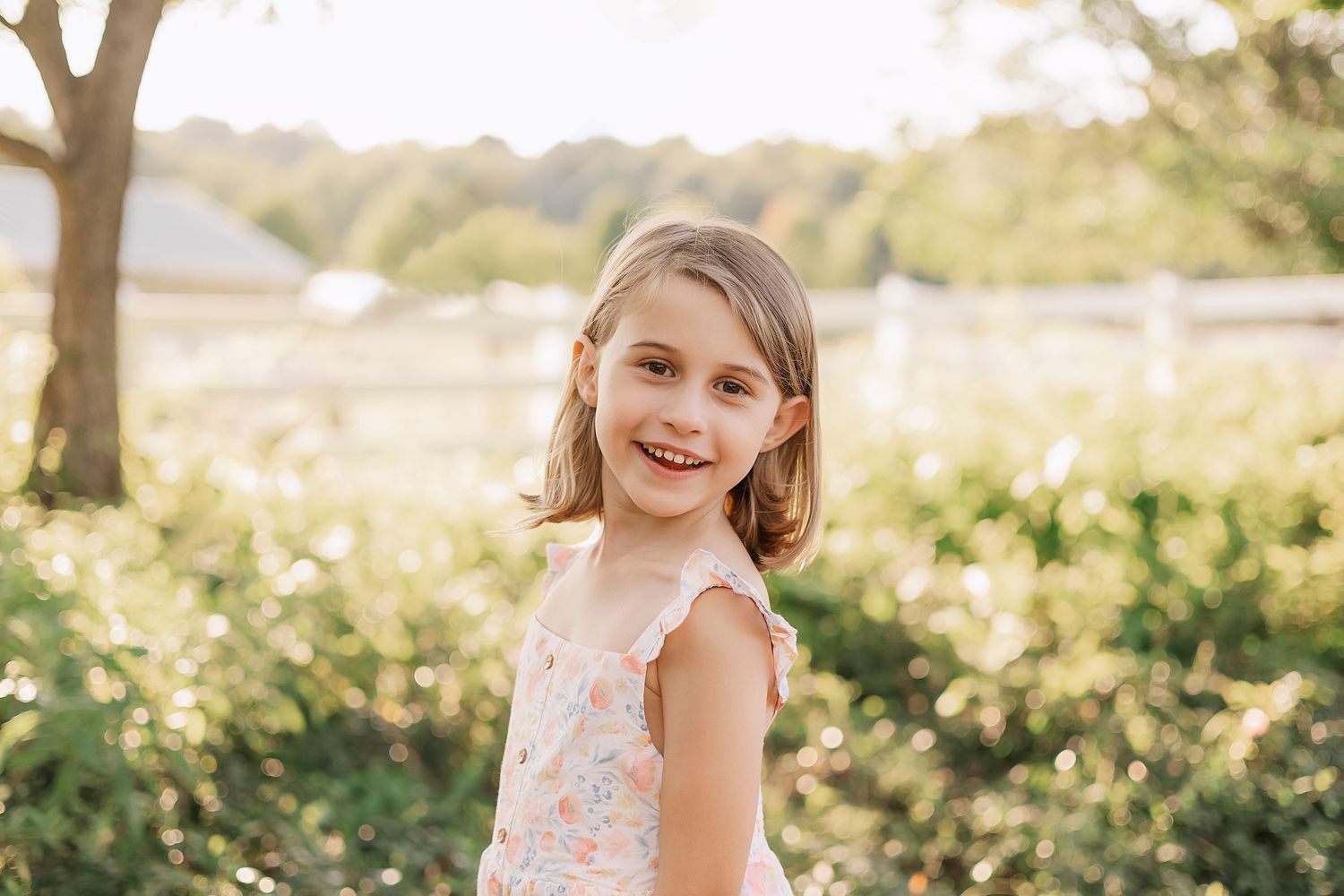 Child in a floral dress smiles in golden sunlight with trees and grass in background.