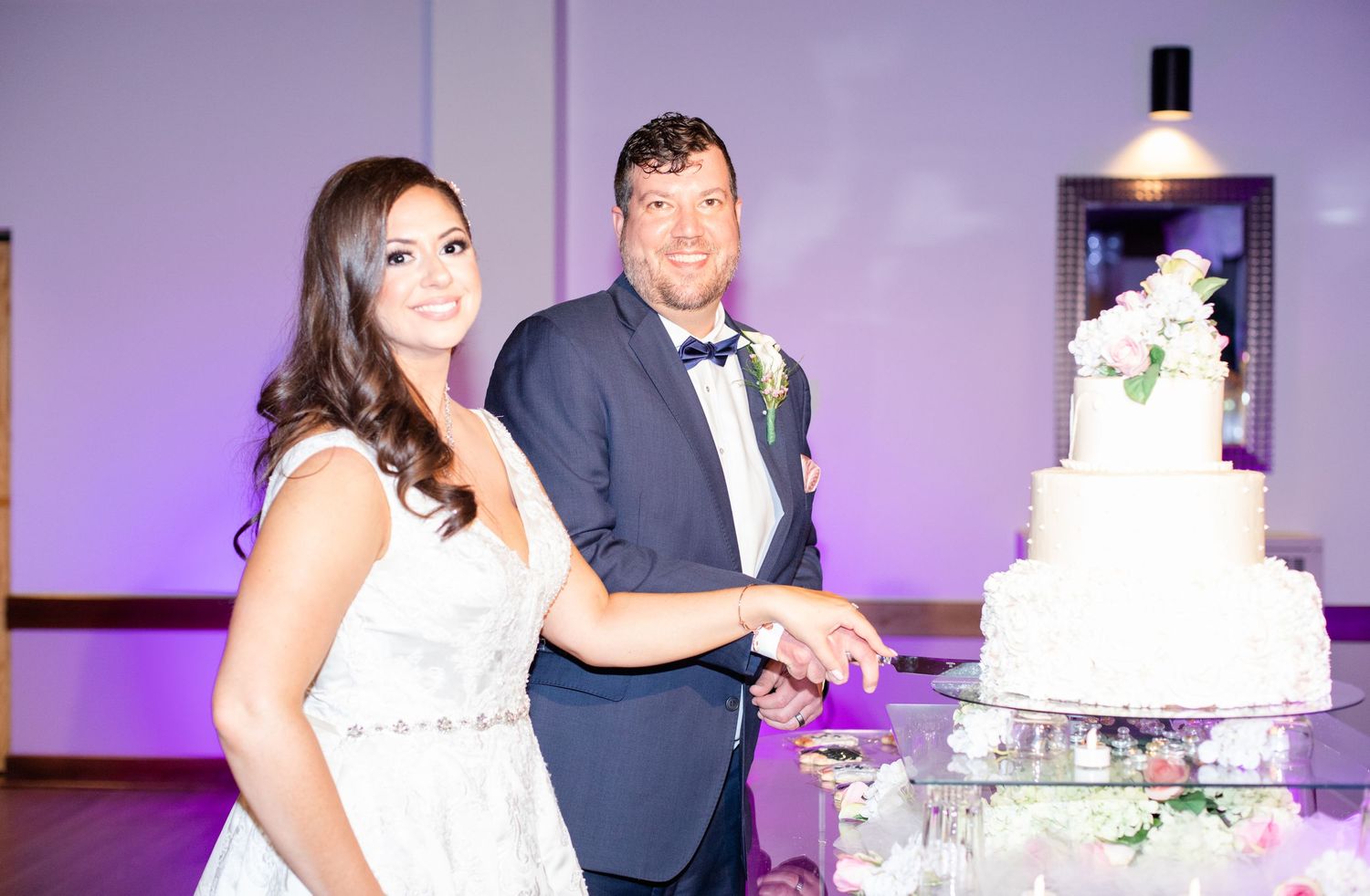 A couple shares a moment while cutting their multi-tiered white wedding cake at their reception.