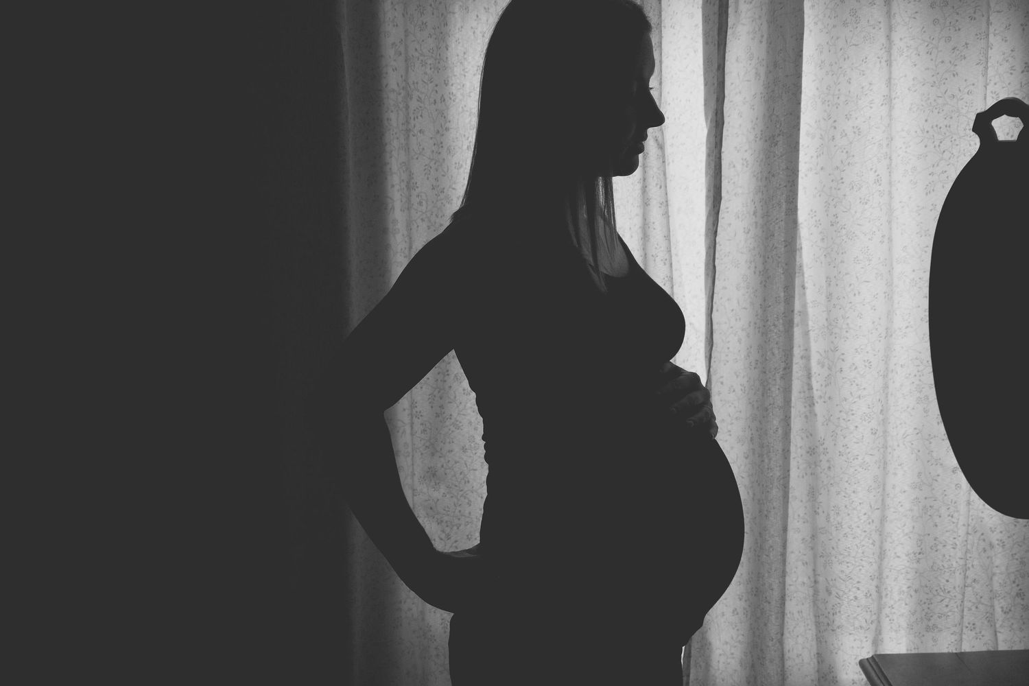 A silhouetted profile of a pregnant woman stands against a window with curtains.