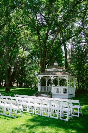 White gazebo set up for outdoor wedding ceremony with rows of chairs under green trees.