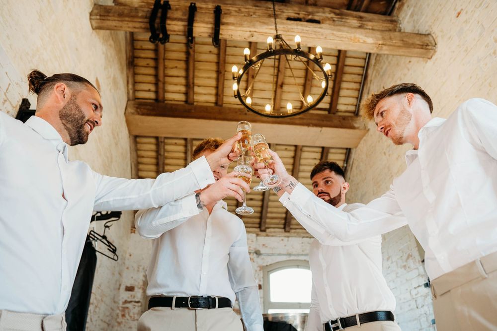 Four men in white shirts toast champagne glasses under a rustic wooden beamed ceiling.