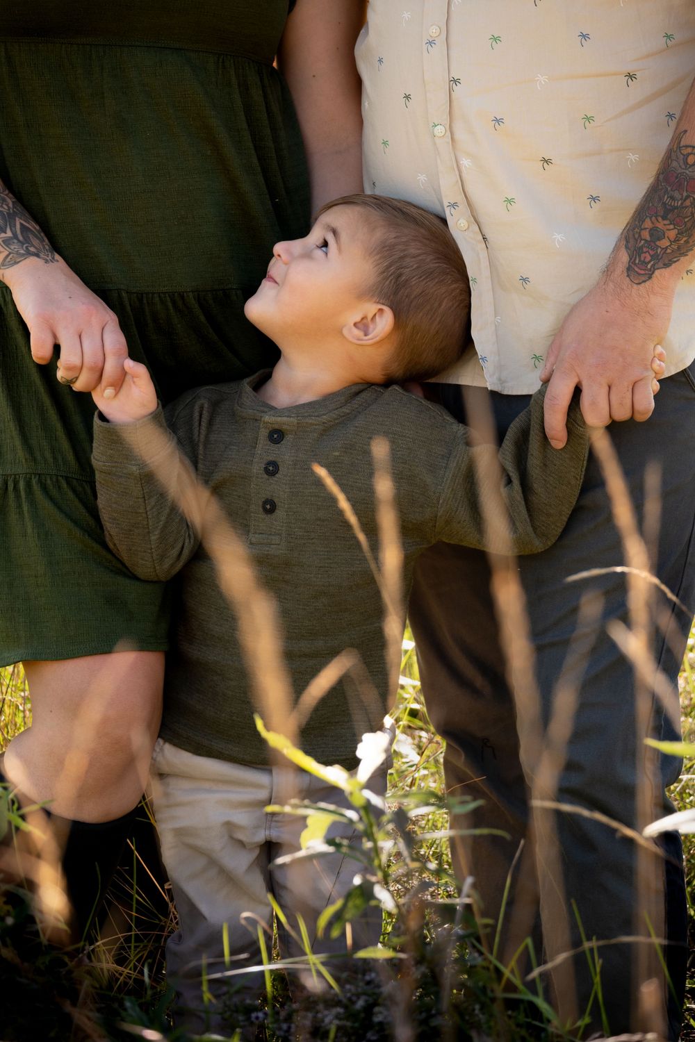 Young boy looks up smiling while standing between parents outdoors in a sunny field.