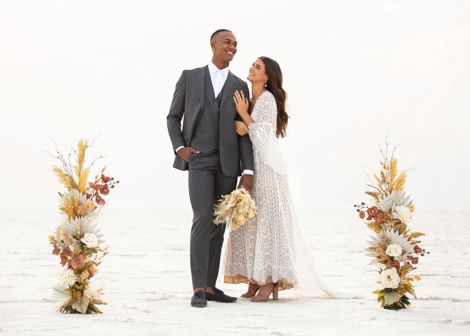 A couple poses for a romantic wedding photo on a white beach with boho floral arrangements on either side.
