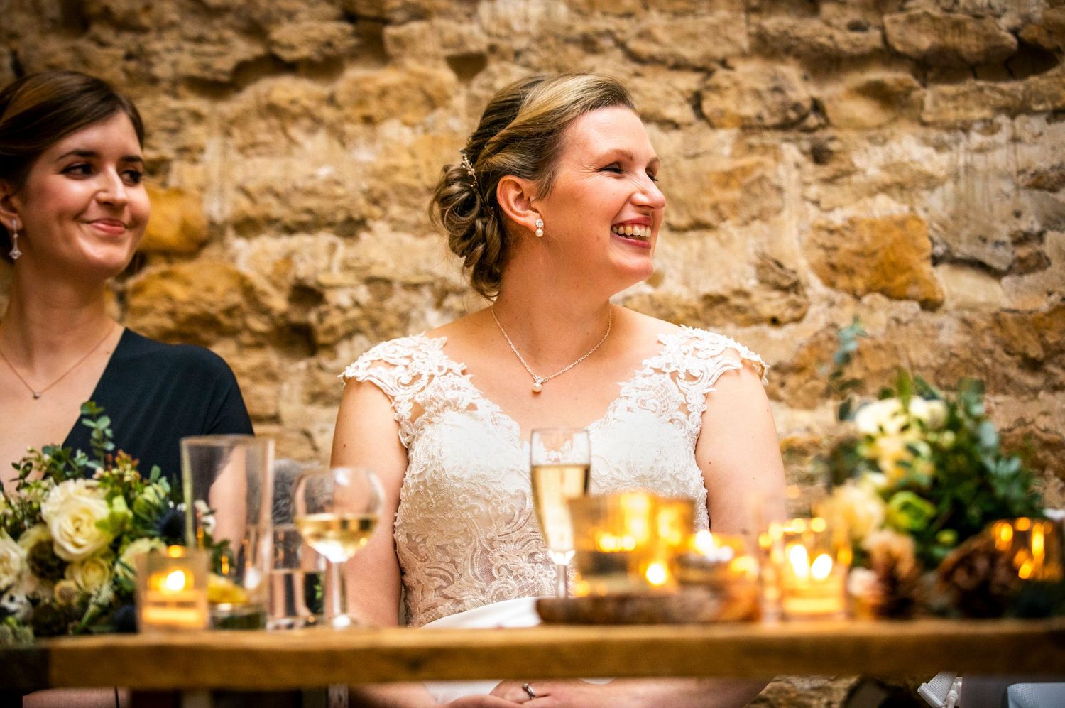 Wedding reception dinner setup against a rustic stone wall with warm candlelight and floral arrangements.
