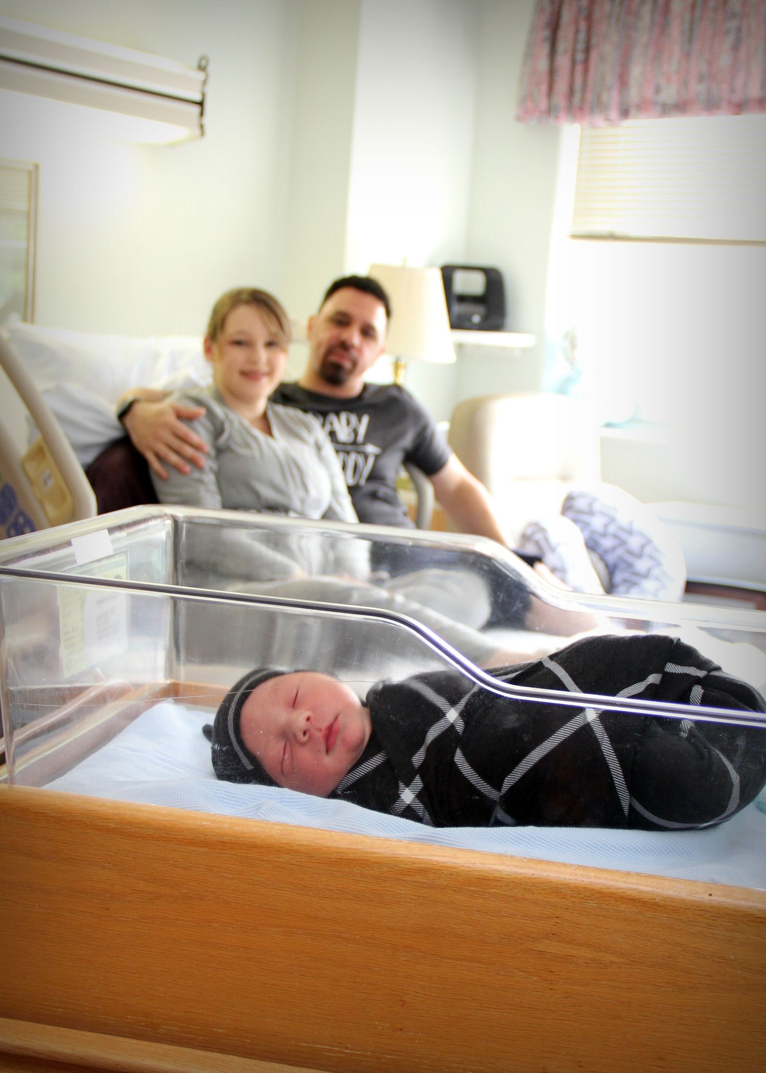 A newborn baby sleeps in a hospital bassinet while parents watch nearby.