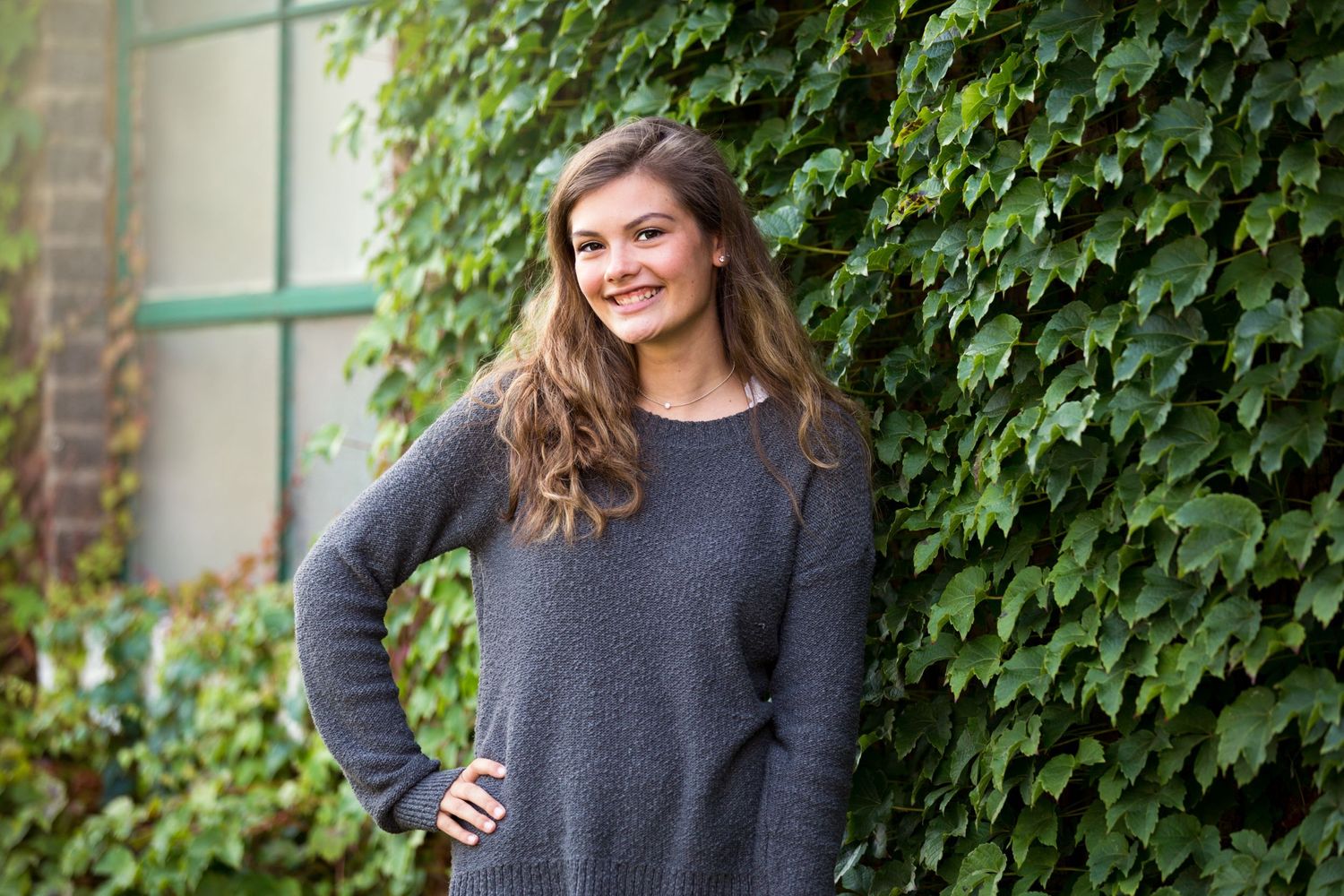 Senior portrait session against ivy covered wall featuring casual gray sweater.