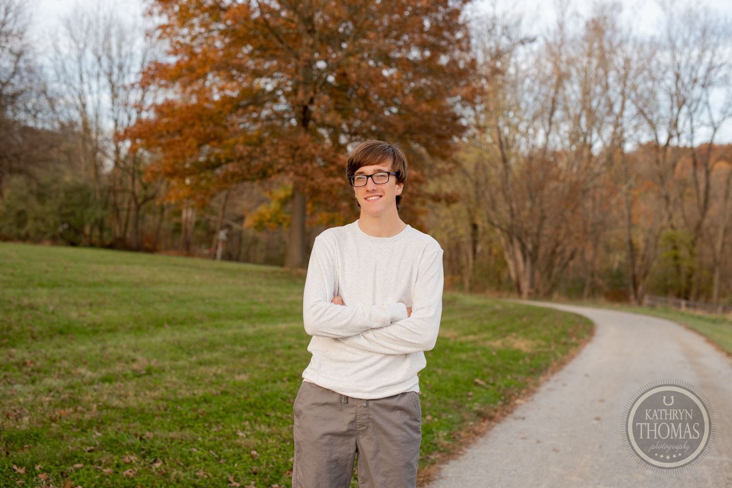 A person in a white sweater stands on a curved path with fall foliage in the background.