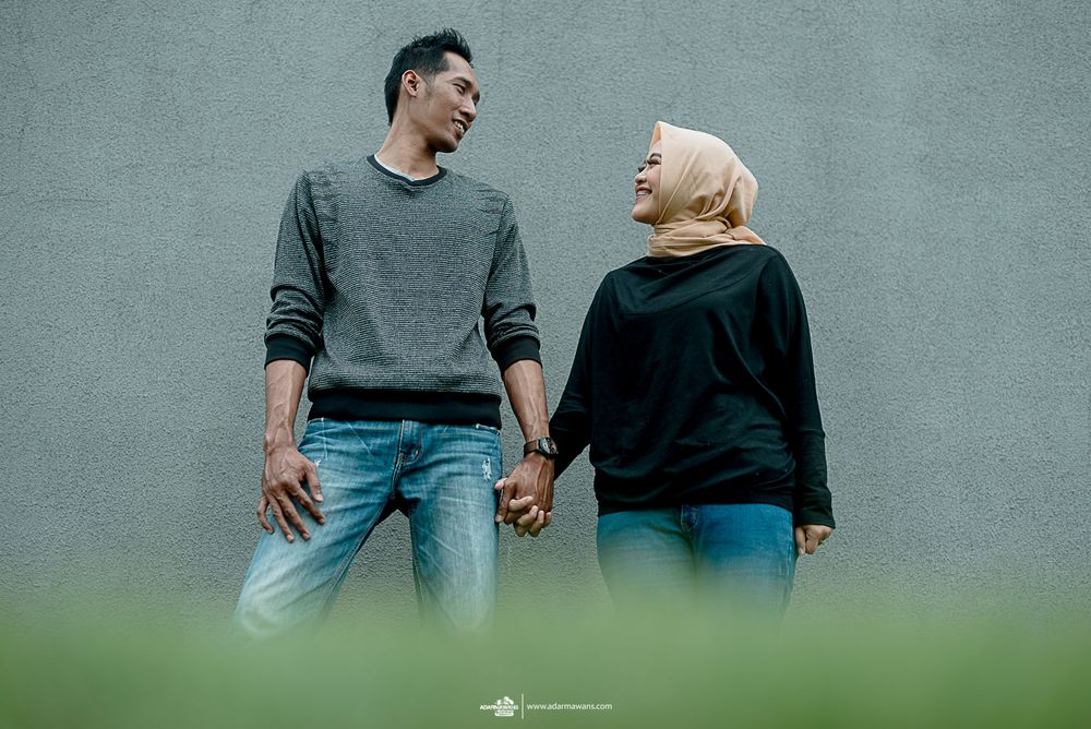 Couple in dark sweaters sharing a laugh against grey wall with green foreground.