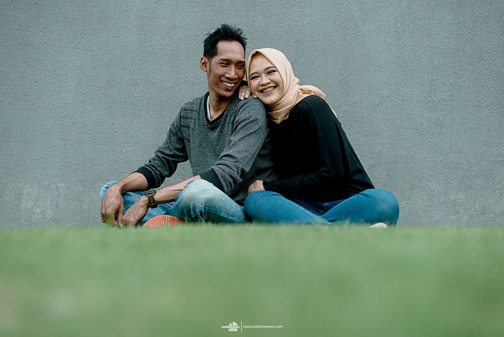 A couple sits together on grass against a gray wall sharing an intimate moment.
