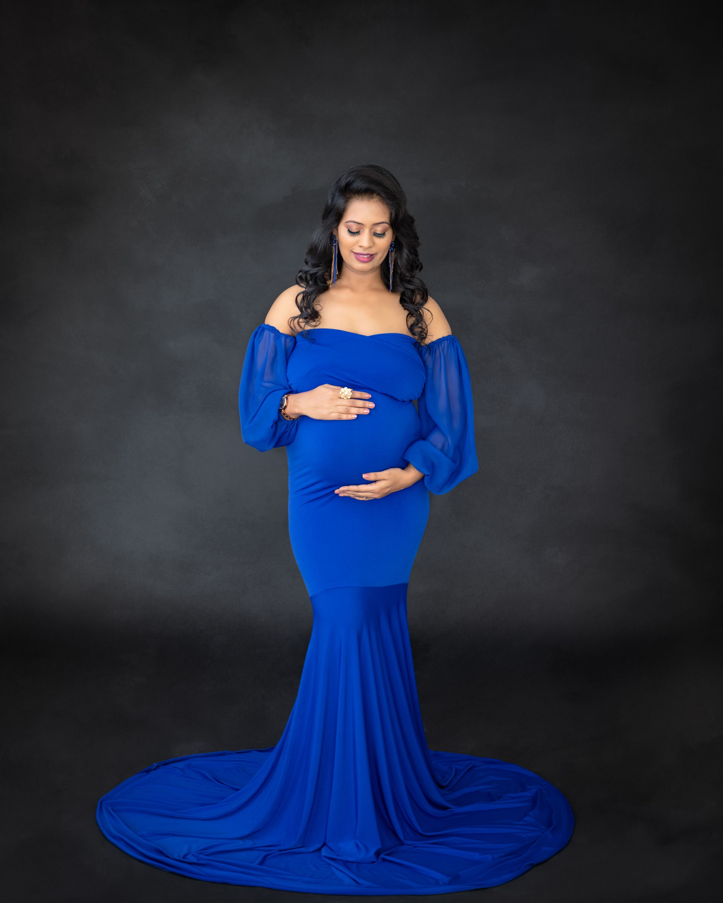 Maternity portrait in royal blue off-shoulder gown with flowing train against dark background.
