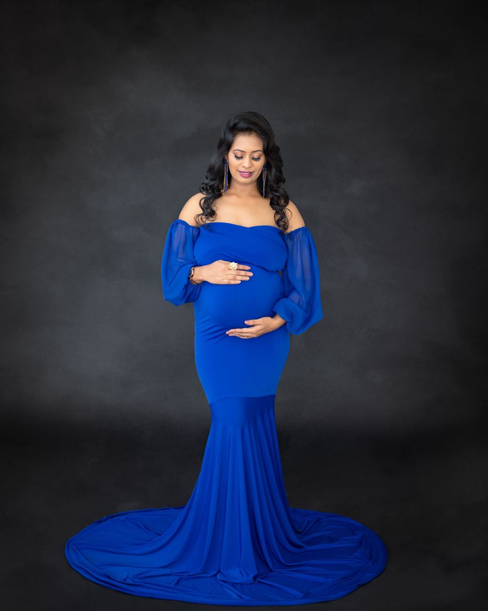 Maternity portrait in royal blue off-shoulder gown with flowing train against dark background.