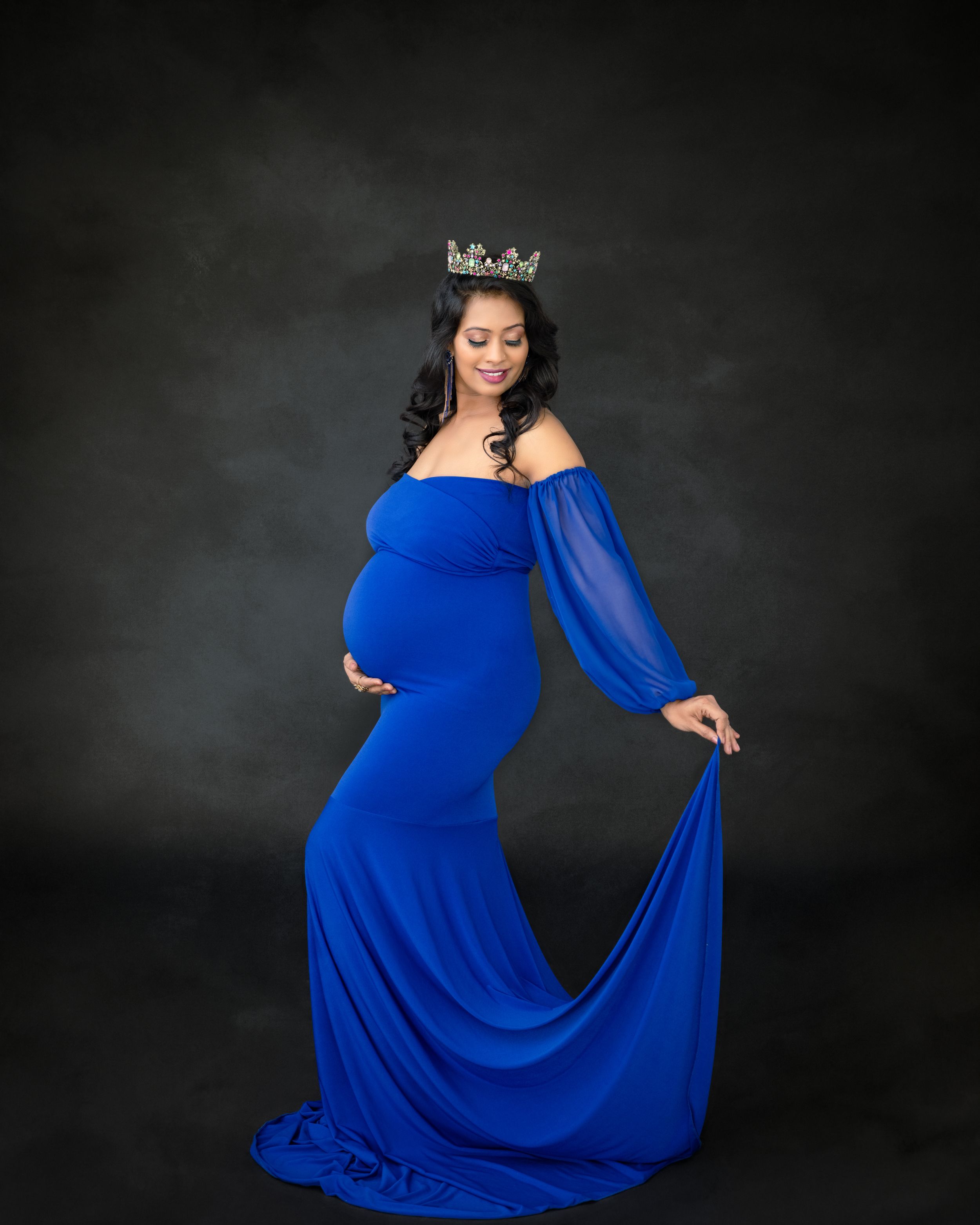 Elegant maternity photo featuring a flowing royal blue dress and crown against dark backdrop.