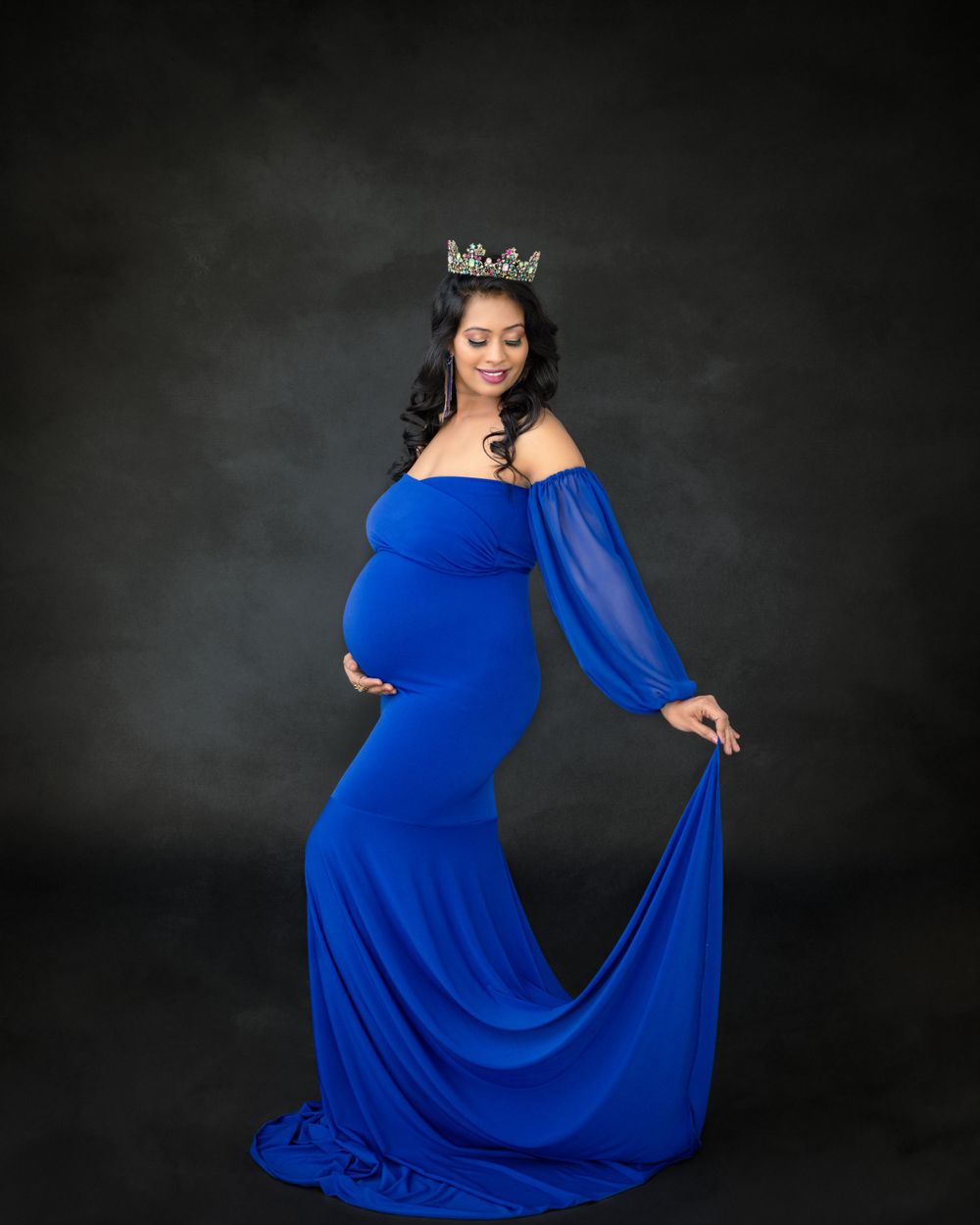Elegant maternity photo featuring a flowing royal blue dress and crown against dark backdrop.