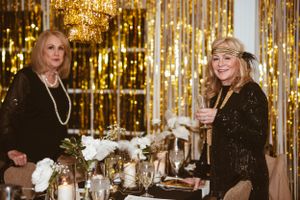 Two guests in formal attire celebrate at an elegant party with gold decor and chandeliers in the background.