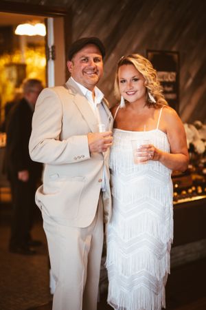 Couple in white formal attire poses with drinks at an elegant party.