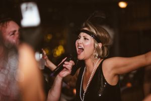 A performer in flapper-style dress and pearls sings passionately into a microphone at a dimly lit party.