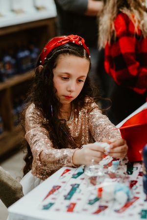 Child in sparkly dress and red headband works on holiday crafts at table.