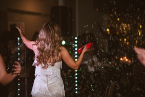 A woman in a white dress dances energetically with raised arms among glittering party lights.