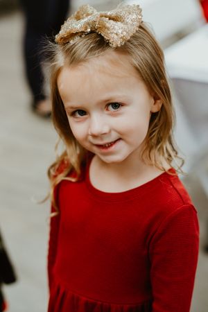 Young child wearing a red sweater and sparkly bow smiles warmly at the camera.