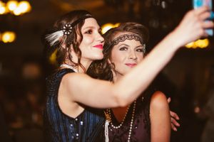 Two partygoers in 1920s flapper style dresses take a selfie together at a glamorous event.
