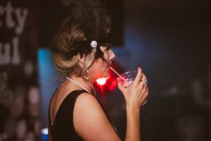 A performer with a feathered headpiece dances energetically under red lighting at a party.