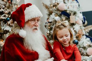 Traditional Santa Claus figure sits with child in red dress near decorated Christmas tree.